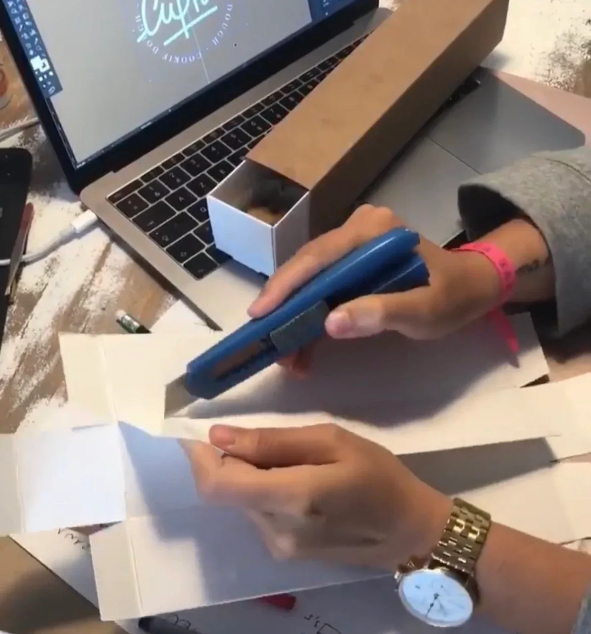 Mains découpant au cutter un prototype de boîte en carton, le logo Cupkie à l’écran d’un ordinateur portable