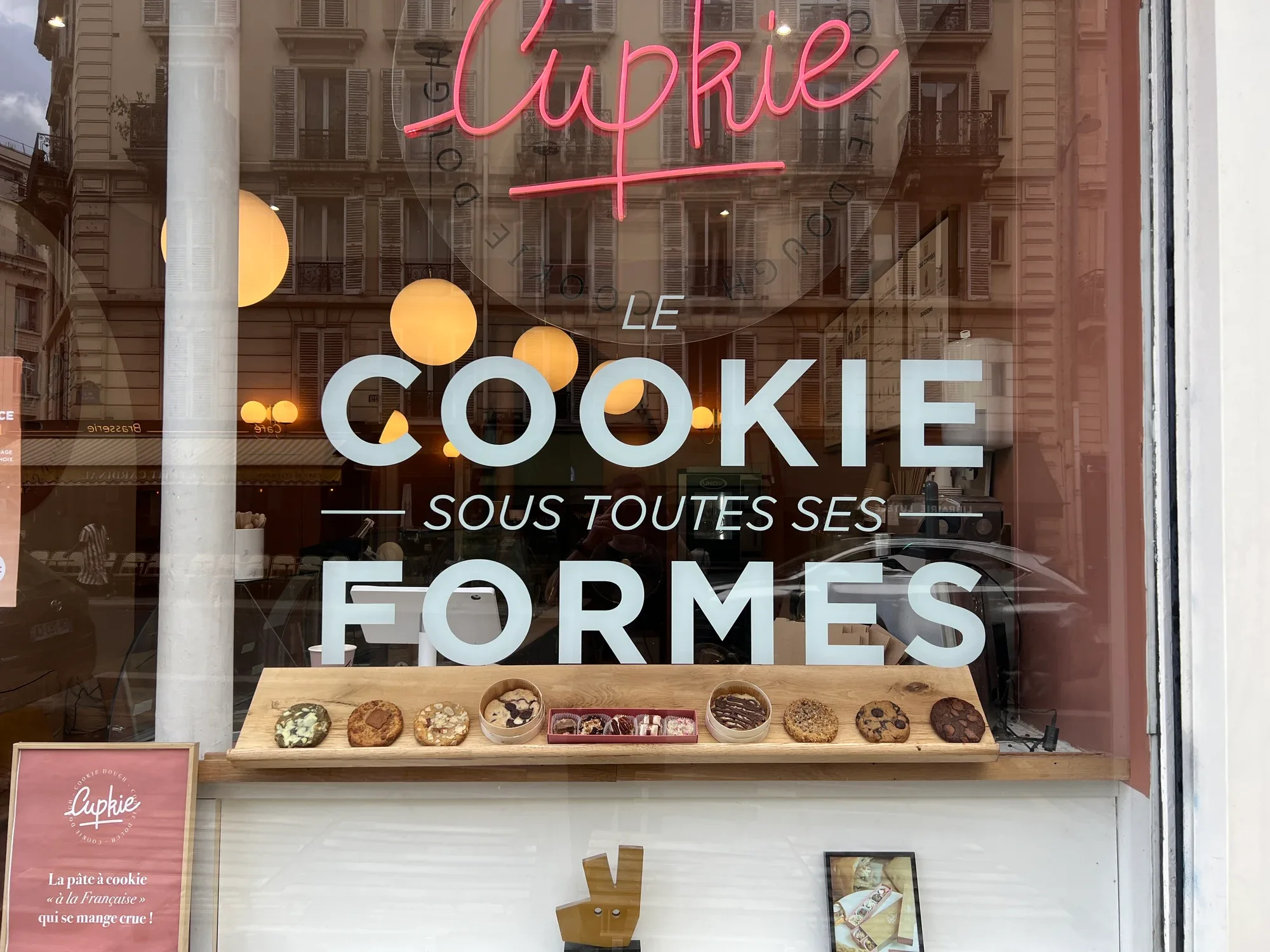 La vitrine Cupkie avec le néon rose et la vitrophanie « Le cookie sous toutes ses formes », cookies alignés sur une planche en bois